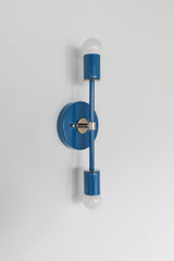 Angled product image of the Medium Thalia Sconce in Denim Blue with chrome accent hardware, shown against a clean white wall. The simple round backplate, vertical arm, and exposed globe bulbs give this blue wall light a fresh modern feel, making it a strong fit for bathroom mirrors, narrow powder rooms, colorful hallways, kids spaces, and retro-inspired interiors.