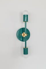 Close angled image of the Mermaid Green Medium Thalia Sconce with brass hardware and two white globe bulbs. The photo shows the fixture’s glossy teal green finish, round wall plate, slim vertical arm, and warm metallic accents. This colorful wall light works well for bathroom vanity lighting, powder room sconces, hallway lighting, bedroom accent lighting, and interiors with coastal, retro, chinoiserie, or grandmillennial style.