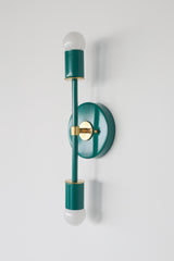 Angled studio photo of the Medium Thalia Sconce in glossy Mermaid Green with warm brass hardware and two white globe bulbs. This colorful vertical wall sconce has a jewel-tone teal green finish that works beautifully in bathroom vanity lighting, powder rooms, kids bathrooms, hallways, bedrooms, and playful interiors with brass accents, vintage-inspired details, modern color, or grandmillennial style.