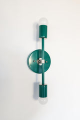 Angled product photo of the Medium Thalia Sconce in Mermaid Green with chrome hardware, shown against a simple white wall. The image highlights the round backplate, slim vertical arm, glossy green powder-coated finish, and two globe bulbs. This two-light wall sconce works beautifully in colorful bathrooms, playful powder rooms, modern vintage interiors, and spaces that need a bright green accent light.
