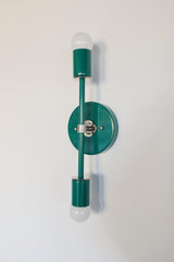 Angled product photo of the Medium Thalia Sconce in Mermaid Green with chrome hardware, shown against a simple white wall. The image highlights the round backplate, slim vertical arm, glossy green powder-coated finish, and two globe bulbs. This two-light wall sconce works beautifully in colorful bathrooms, playful powder rooms, modern vintage interiors, and spaces that need a bright green accent light.