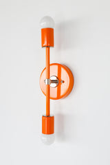 Angled view of the Medium Thalia wall sconce in glossy orange peel with chrome hardware and visible round backplate depth, modern sculptural vertical double globe sconce with two white globe bulbs and powder coated steel finish, bold color pop wall lighting for bathroom vanity lighting, hallway wall sconces, bedside reading light, eclectic modern and maximalist decor, designer statement lighting handmade by Sazerac Stitches