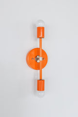 Angled view of the Medium Thalia wall sconce in glossy orange peel with chrome hardware and visible round backplate depth, modern sculptural vertical double globe sconce with two white globe bulbs and powder coated steel finish, bold color pop wall lighting for bathroom vanity lighting, hallway wall sconces, bedside reading light, eclectic modern and maximalist decor, designer statement lighting handmade by Sazerac Stitches