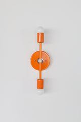 Front view of the Medium Thalia wall sconce in bright orange peel with polished chrome hardware, a modern mid century inspired vertical double globe wall light with round backplate and two white globe bulbs, bold colorful statement sconce for bathroom vanity lighting, hallway sconces, bedside wall lighting, kitchen accent light, dopamine decor and maximalist interior design, handmade by Sazerac Stitches