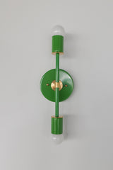Front-facing studio photo of the Medium Thalia Sconce in glossy Spring Green with warm brass hardware and two white globe bulbs. This colorful vertical wall sconce has a clean, playful shape that works beautifully for bathroom vanity lighting, kids bathrooms, powder rooms, hallways, bedrooms, and cheerful interiors that need a bright pop of green.