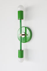 Side-angle studio image of the Medium Thalia Sconce in Spring Green with chrome hardware, shown on a clean white wall with two white globe bulbs. The saturated green finish gives this wall light a bold but simple look, perfect for colorful bathroom lighting, kids rooms, powder rooms, bedrooms, and fresh modern or eclectic interiors.