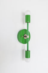 Angled product photo of the Medium Thalia Sconce in glossy Spring Green with chrome accent hardware. The slim two-light design gives the fixture a narrow profile while still adding strong color, making it a fun option for bathroom mirrors, small powder rooms, colorful kids spaces, hallways, and modern interiors with a playful edge.