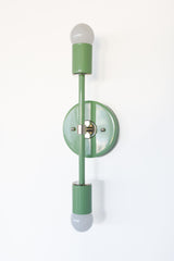 Side angle view of the Medium Thalia wall sconce in vista green with chrome hardware, featuring a slim vertical stem and double bulb design, a modern mid century lighting fixture made for bold interiors, bathrooms, and playful patterned wallpaper.