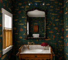 Dark green fish and floral wallpaper sets the tone in this traditional powder room with an antique wood vanity, marble counter, brass faucet, mustard café curtain, and ornate vine-framed mirror. The blue Medium Thalia Sconce above the mirror adds a clean modern note, with a round backplate, slim horizontal arm, brass accents, and white globe bulbs.