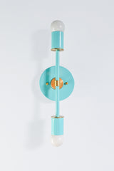 Glossy turquoise metal with brass hardware and two bare globe bulbs. The Medium Thalia Sconce is a vertical bar mounted on a round backplate, with a brass collar at the center and brass discs at the base of each socket. The Poolside color is bright and clean, and the brass keeps it from feeling too sweet. Try a pair in a bathroom, a bedroom or a hallway that needs a little color.
