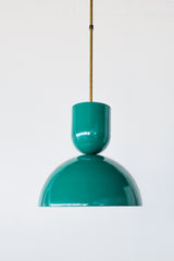 Deep emerald-green is the color that makes a kitchen feel rich and a little jewel-like, and this Taeuber pendant brings it in a punchy, sculptural shape. The two-part silhouette stacks a rounded cup over a wide domed bell, all finished in a glossy mermaid green with a warm brass stem, shown here lit and glowing against white brick. It reads saturated and confident, the kind of statement lighting that turns an island, breakfast nook, or sink into a focal point.