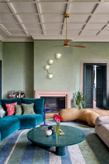 Maximalist living room photo featuring the Aries wall sconce in a mint green finish, a sculptural modern wall light with four round disc shades and white globe bulbs arranged in a curved, constellation-like layout above a pale pink fireplace mantel. The fixture pops against soft sage green walls and a classic white coffered ceiling, styled with a brass-and-wood ceiling fan, teal velvet sectional sofa, green stone coffee table, layered patterned rug, and eclectic decor accents. 