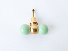 A double-ball drawer pull pairing two soft mint green spheres with a polished brass ball at the center, shown here with a gentler, more matte finish. A quiet pastel pick for a nursery, a soft kitchen palette, or bathroom cabinetry that wants color without going bold.