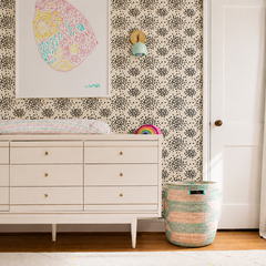 Close-up nursery styling: a mint green Loa globe wall sconce with brass backplate mounted on black and white dot floral wallpaper above a white midcentury modern dresser and changing pad. A colorful abstract art print and woven storage hamper add playful texture, making this a Pinterest-friendly modern nursery lighting idea.