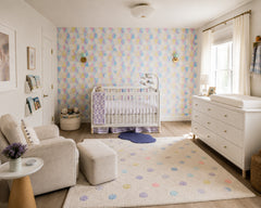 Playful modern nursery with pastel triangle wallpaper and a pair of mint green Loa wall sconces in brass, flanking a white crib. Soft, kid-friendly styling includes a cozy upholstered rocker, white mid century dresser, dotted area rug, and natural light. Ideal Pinterest inspo for nursery wall lighting, mint sconce, brass nursery sconce, and colorful Scandinavian nursery decor.