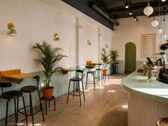 Sunlit modern cafe interior with a row of mint green Loa wall sconces in brass along a white wall with sage wainscoting. Minimalist cafe seating, black stools, warm wood tables, potted palms, and a mint countertop create a fresh mid century vibe. Search-friendly inspiration for cafe wall lighting, restaurant sconces, brass wall sconces, and minimalist commercial interior design.