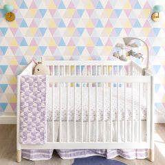 Close-up of a pastel geometric accent wall behind a white crib, styled with two mint green Loa wall sconces in brass facing upward. Soft triangle wallpaper in pink, blue, yellow, and lavender pairs with a purple quilt and modern baby mobile. Ideal for nursery lighting inspiration, mid century nursery sconce, mint green wall light, and colorful kids room decor.