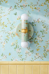 A close-up of the Mint, Cream, and Gold Marbled Small Thalia Sconce styled on light blue floral wallpaper with white blossoms, birds, butterflies, and butter yellow tile wainscoting. The handmade marbled finish adds a one-of-a-kind decorative detail while the small scale makes it ideal for bathroom vanity lighting, powder rooms, children’s bathrooms, and colorful cottage-inspired spaces.