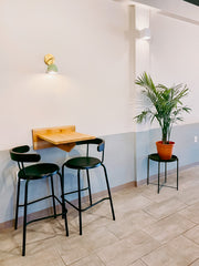 Minimalist restaurant nook with a mint green Loa wall sconce in brass above a compact wood counter table and black cafe chairs. Clean white walls with soft sage wainscoting, tiled floor, and a potted palm give it a fresh modern vibe. Great for commercial lighting inspiration, cafe sconce, brass wall lighting, and small space dining ideas.