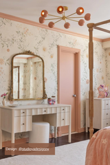 Elegant bedroom interior featuring a brass and peach mid-century modern chandelier, floral wallpaper, soft peach-painted trim and door, a vintage-inspired gold mirror, scalloped wooden vanity table with upholstered stool, and tasteful decorative accents creating a soft, feminine aesthetic.