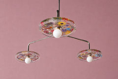 Studio photo of the Botanico chandelier with chrome arms, white globe bulbs, and clear circular shades filled with colorful botanical flowers. The mauve pink background makes the floral pattern feel playful, feminine, and artful, while the chrome hardware keeps it crisp and modern. A statement ceiling light for maximalist interiors, dopamine decor, garden-inspired rooms, colorful kitchens, creative offices, nurseries, kids spaces, and eclectic homes that need a floral chandelier with personality.