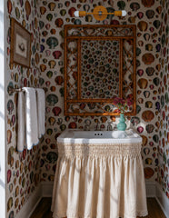 Coastal cottage bathroom with colorful shell wallpaper, a skirted white sink, bamboo mirror, turquoise vase, and warm natural light. The Mustard and Chrome Thalia Sconce sits above the mirror with a round mustard backplate, matching cylindrical socket covers, chrome arm, and white globe bulbs. The palette mixes mustard yellow, cream, shell pink, brown, green, and silver chrome.
