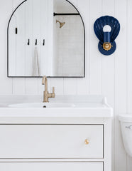 Clean neutral bathroom with white vertical shiplap, a black arched mirror, simple white vanity, brushed metal faucet, and a single Navy Coquille sconce mounted beside the mirror. The shell-shaped blue wall light adds sculptural color to an otherwise minimal bathroom, making it feel coastal, modern, and a little more collected. This photo fits navy wall sconce, shell bathroom sconce, coastal bathroom lighting, small bathroom vanity light, neutral powder room, modern farmhouse bathroom, and classic blue and w