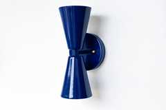 A statement wall sconce for rooms that lead with saturated color and sculptural shapes. This angle of the Dryades sconce shows off its glossy navy double-cone silhouette — one cone throwing light up, one down — meeting at a narrow waist on a round navy backplate. Deep, graphic, and a little Art Deco, it's a confident pick for hallways, bars, bedrooms, and colorful traditional or mid-century interiors that want bold color without losing polish.