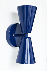 A bold double-cone wall sconce for a hallway, bar, bedroom, or mid-century modern room that wants saturated color and crisp, two-directional light. The Dryades sconce pairs an upward and a downward glossy navy cone at a narrow waist, mounted on a round navy backplate with a brass screw accent. Seen from an angle, its bowtie silhouette and wall projection read graphic and confident — a classic-but-daring pick for colorful traditional, coastal, and modern eclectic spaces.