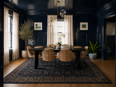Moody, color-drenched navy dining room with high-gloss navy walls and ceiling, paired with a patterned navy area rug and warm wood floors. A brass and clear glass globe chandelier (Newcomb style) creates a modern statement over a long black dining table set with natural woven rattan dining chairs. Soft cream curtains filter daylight at tall windows, while potted greenery adds an organic, coastal-modern touch. Perfect inspiration for dramatic blue dining rooms, transitional interiors, and elevated modern tra