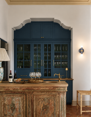 A navy, cream, and gold marbled Thalia wall sconce with raw brass hardware is switched on, casting a warm glow on a white wall next to a deep navy, glass-front built-in cabinet. In the foreground, a rustic, vintage-style wood bar island holds wine bottles, a tray of stemware, and a brass bar faucet.
