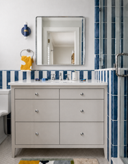 A blue hand-marbled Thalia wall sconce with brass detailing is shown in a clean blue and white bathroom beside a chrome-framed mirror. The space features glossy blue striped tile, a white vanity, chrome hardware, and a playful yellow towel for a fresh, slightly coastal look. This image helps show how a blue marbled wall sconce can add color and personality to a bathroom without overwhelming the space.