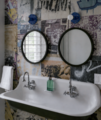 This shared bathroom pairs a long cast iron trough sink with two separate faucets and two round black-framed mirrors, all set against a dense, poster-and-graffiti collage wallpaper. A pair of navy and silver Sid Sconces sits above each mirror, their spiked domes adding an industrial edge that fits the wallpaper's downtown energy. It's a smart layout for siblings or roommates who need their own space at the same sink.

