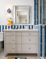 This image shows a blue marbled Thalia sconce illuminated beside a rectangular mirror in a bright bathroom with blue striped tile, a white vanity, and chrome accents. The warm glow softens the crisp blue-and-white palette and highlights the sconce’s handmade marbled finish and brass center detail. It’s a great example of a colorful bathroom wall light with a playful coastal feel.