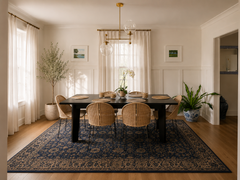 Airy, family friendly dining space styled in a soft neutral palette with white paneled walls, warm oak floors, and breezy sheer curtains. A brass Newcomb chandelier with oversized clear globe lights adds modern statement lighting above a matte black dining table. Rattan cane chairs bring coastal texture, paired with a navy blue patterned area rug for contrast. Green plants in blue and white ceramic planters and simple wall art finish the relaxed transitional, modern classic dining room design.
