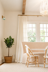 Bright neutral dining nook featuring a mint green Loa wall sconce with brass backplate and arm, mounted beside tall linen curtains and sunlit windows. Soft coastal-modern styling with a woven pendant light, warm wood ceiling beam, rattan bistro chairs, and a potted plant on a textured rug. Great inspiration for mid century modern wall lighting, brass sconces, and airy dining room decor.