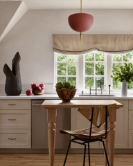 See the handmade terracotta Alexander Pendant styled in a soft neutral kitchen — its matte, clay-toned dome and brass finial adding a warm earthy anchor above a white oak table, linen valance, and marble counters. This is quiet color done right: a muted terracotta light fixture that brings just enough warmth to a calm, organic-modern space full of natural wood, stone, and greenery. Perfect inspiration for a breakfast nook, cottage kitchen, or warm-minimalist remodel. Shop handmade terracotta pendant lightin