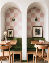 Bright restaurant nook with twin arched booths, pink and cream checker wall panels, olive green channel-tufted banquettes, natural wood tables, and matching wood dining chairs. The Olive and Brass Adele sconces add small-scale statement lighting above each booth, bringing a playful mix of modern cafe design, retro diner charm, and soft maximalist color. This photo works well for restaurant lighting, cafe booth lighting, olive green wall sconces, checkerboard interiors, hospitality design, and colorful banqu