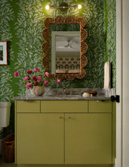 Lit Olive and chrome Medium Thalia Sconce installed as a horizontal vanity light above a scalloped wicker mirror in a richly patterned green bathroom. The warm exposed bulbs add glow against leafy green wallpaper, an olive vanity, marble counter, woven accents, and vintage-inspired bathroom styling. A great lighting idea for small powder rooms, guest bathrooms, botanical wallpaper bathrooms, green bathroom designs, cottagecore interiors, and colorful custom vanity lights.