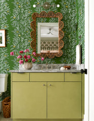Olive and chrome Medium Thalia Sconce shown horizontally above a bathroom vanity mirror in a green tonal powder room. The space features leafy botanical wallpaper, an olive green vanity, marble countertop, scalloped wicker mirror, brass faucet, pink flowers, and layered cottage-style decor. A colorful two-light wall sconce for green bathrooms, botanical powder rooms, grandmillennial interiors, garden-inspired bathrooms, cottage bathrooms, and playful vanity lighting.