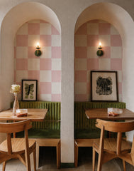 Warm, moody view of a small restaurant or cafe with two arched dining nooks, pink checker walls, deep olive banquette seating, and wood tables. The Olive and Brass Adele sconces are lit above each booth, adding a soft glow that feels intimate, playful, and design-forward without overwhelming the space. A strong fit for boutique restaurant lighting, cozy cafe interiors, modern bistro decor, retro checkerboard walls, green velvet banquettes, and colorful hospitality design.