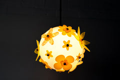 Lit floral acrylic globe pendant against a charcoal backdrop, side profile showing dimensional stacked petals in mixed yellows and chartreuse with subtle transparency, warm ambient glow and sculptural silhouette; single-edition piece designed and made in New Orleans.