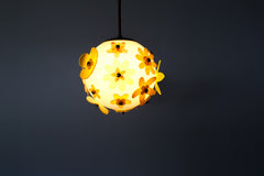 Lit floral acrylic globe pendant against a charcoal backdrop, side profile showing dimensional stacked petals in mixed yellows and chartreuse with subtle transparency, warm ambient glow and sculptural silhouette; single-edition piece designed and made in New Orleans.