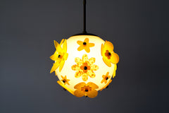 Close view of floral acrylic globe pendant showing layered five-petal flowers in translucent chartreuse and amber with marbled centers, white satin globe diffusion and slim brass stem; single-edition statement light, studio made in New Orleans.