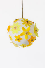 Close view of floral acrylic globe pendant showing layered five-petal flowers in translucent chartreuse and amber with marbled centers, white satin globe diffusion and slim brass stem; single-edition statement light, studio made in New Orleans.