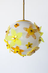One-of-a-kind floral acrylic globe pendant light on white background, white satin-diffuse sphere wrapped in layered yellow and chartreuse acrylic flowers with warm metal caps, suspended from a warm brass stem; cheerful sculptural statement made in New Orleans