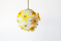 “Close view of floral acrylic globe pendant showing layered five-petal flowers in translucent chartreuse and amber with marbled centers, white satin globe diffusion and slim brass stem; single-edition statement light, studio made in New Orleans