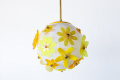 Floral acrylic globe pendant illuminated in a dark room, globe glowing honey warm so petals read like stained glass, layered yellow blossoms and metal caps casting soft halos; unique one-of-a-kind art light by Sazerac Stitches.