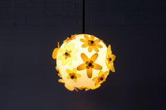 One-of-a-kind floral acrylic globe pendant light on white background, white satin-diffuse sphere wrapped in layered yellow and chartreuse acrylic flowers with warm metal caps, suspended from a warm brass stem; cheerful sculptural statement made in New Orleans