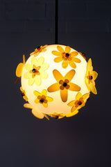 Lit floral acrylic globe pendant against a charcoal backdrop, side profile showing dimensional stacked petals in mixed yellows and chartreuse with subtle transparency, warm ambient glow and sculptural silhouette; single-edition piece designed and made in New Orleans.
